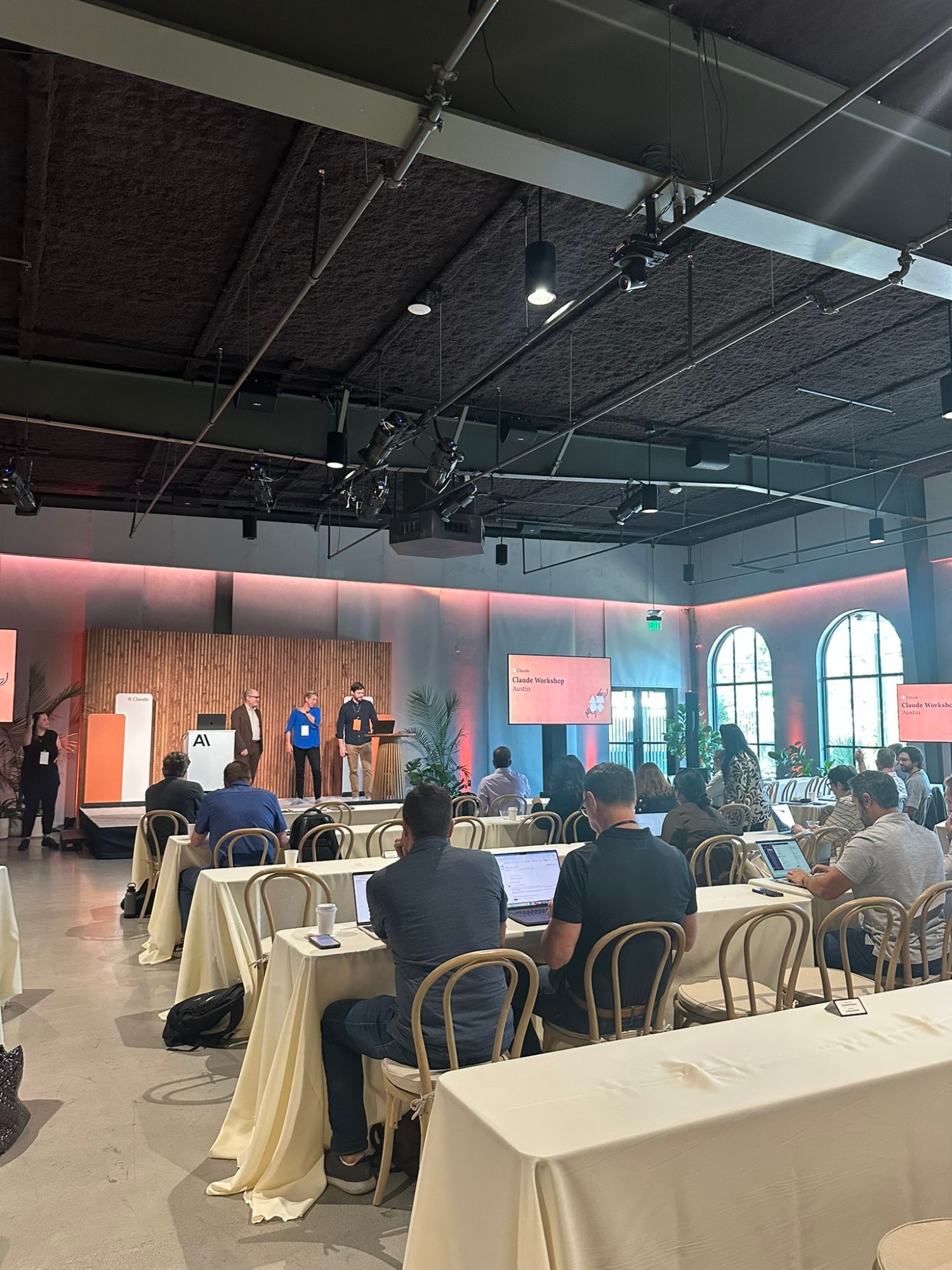Attendees on laptops at long tables during a Claude workshop in Austin, presenters on stage